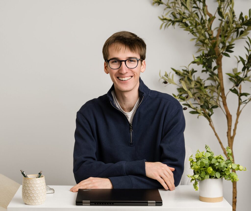 Young man wearing glasses and a dark shirt, smiling at the camera against a plain background