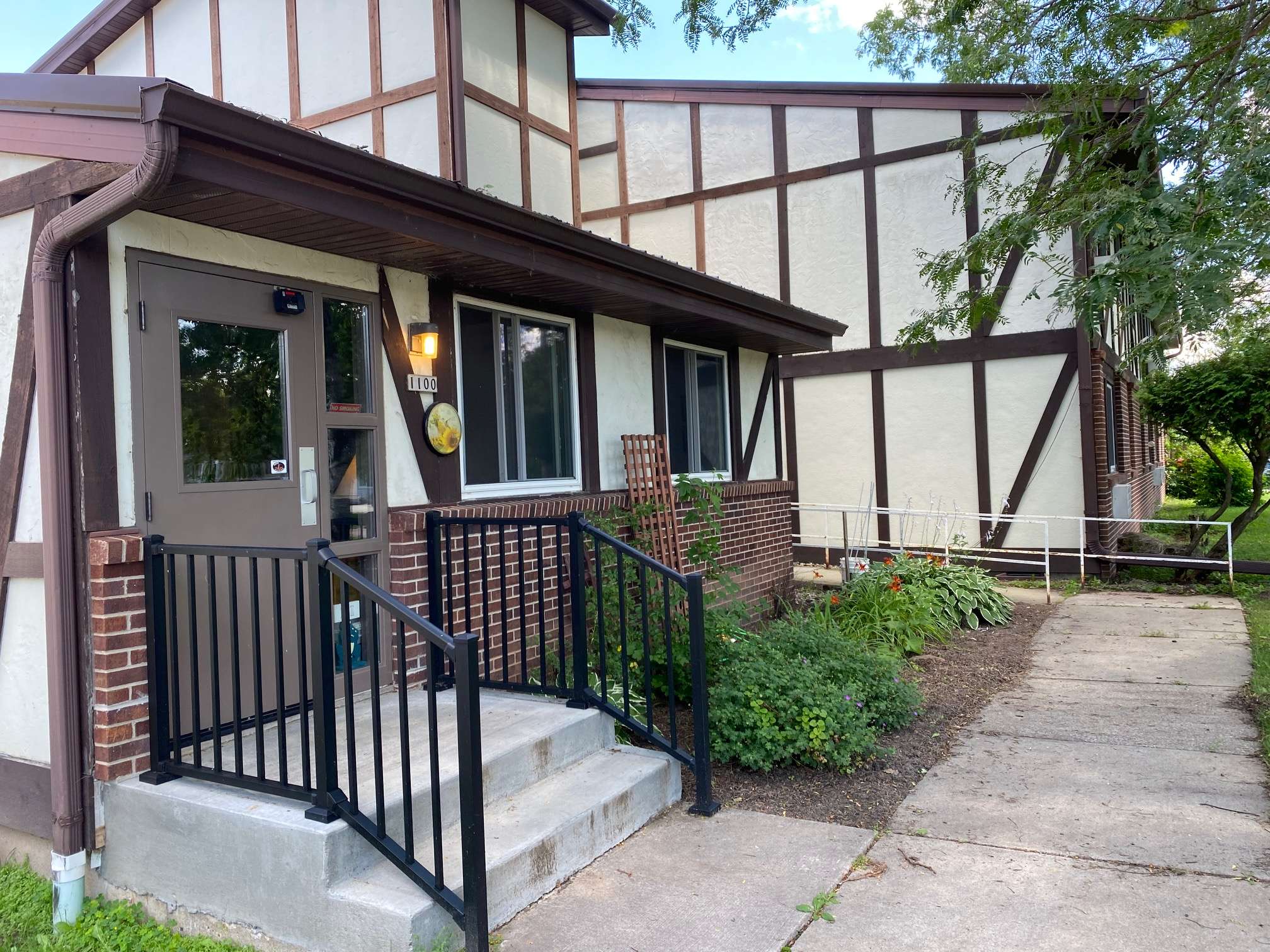 Entryway of a two-story building with a Tudor-style facade, featuring a brown door and adjacent flowerbeds. It showcases accessibility steps and railings.