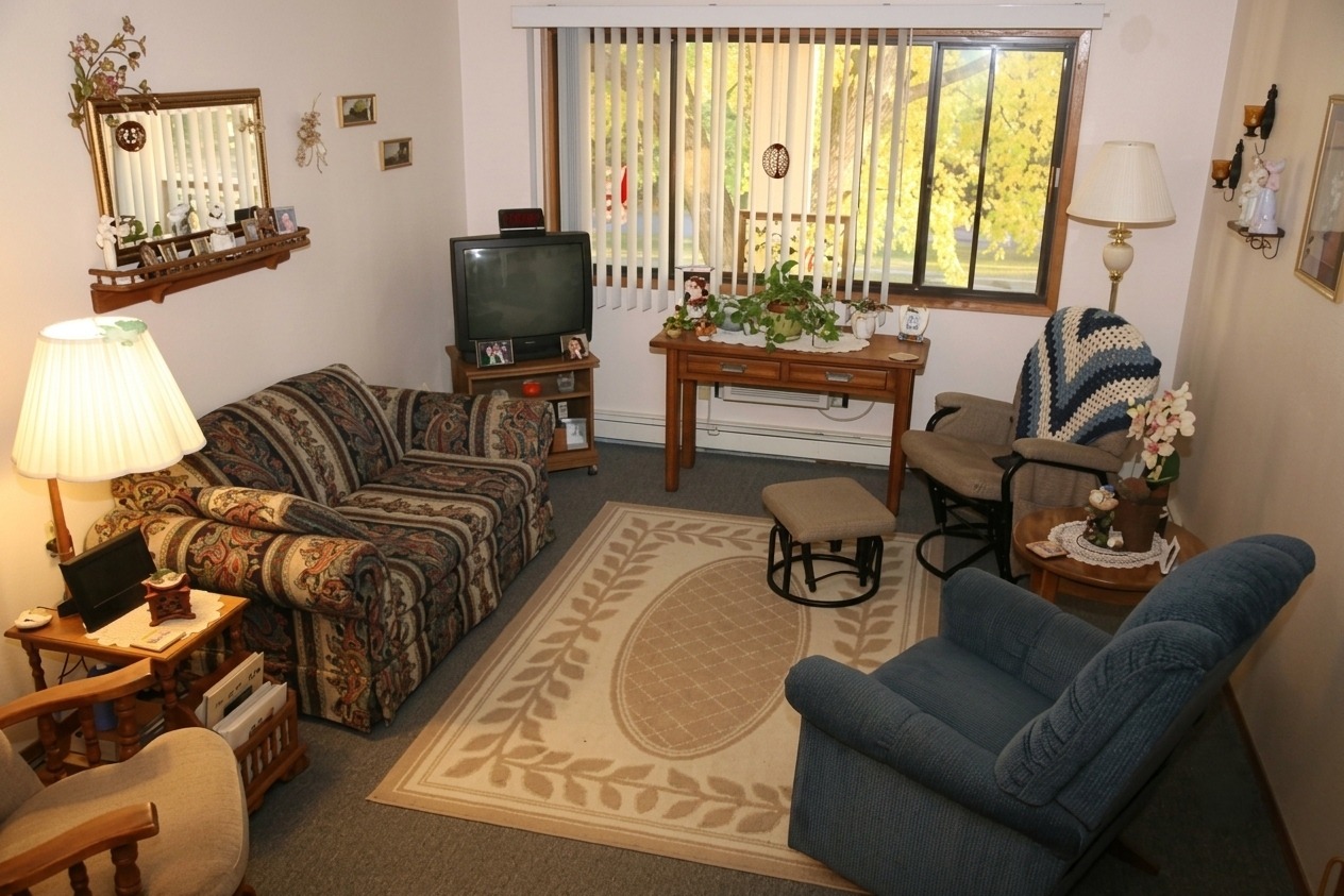 Cozy living room featuring a patterned couch, recliner, and wooden furniture. A window offers natural light, illuminating decor and plants.