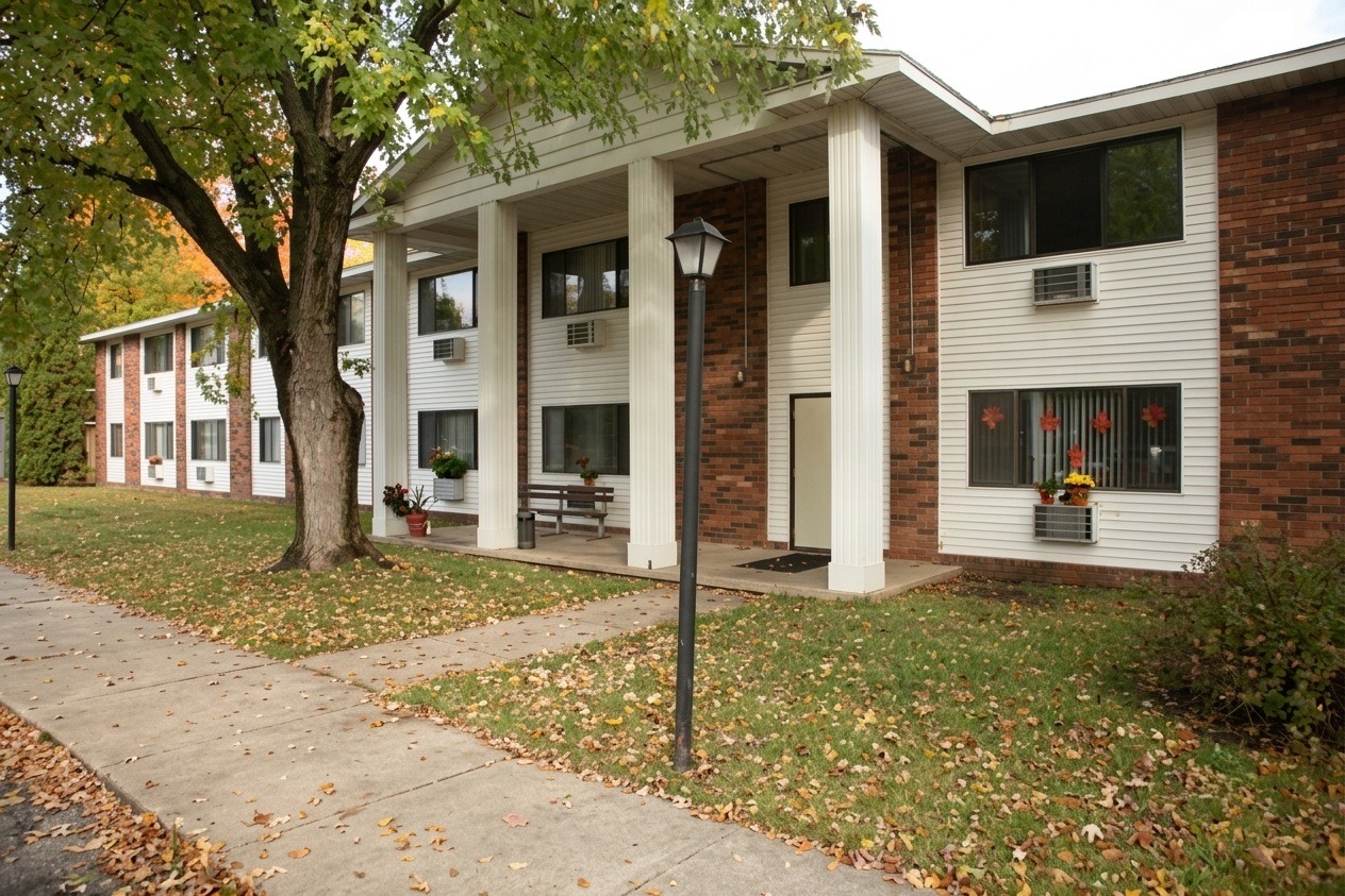 Two-story apartment building featuring brick and siding, surrounded by a grassy area with fallen leaves. A tree and lamp post are visible in front.